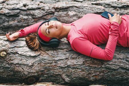 Young happy female runner lying on a wooden log and taking a break with music after running in forest.の写真素材