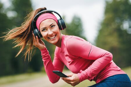 Young cute happy female runner warming up before jogging at morning in forest. She is listening to music on headphones and having fun.の写真素材