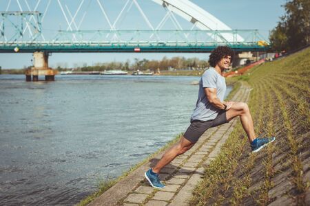 Fit muscular young male runner doing core stretching exercise against a wall, near the river.の写真素材