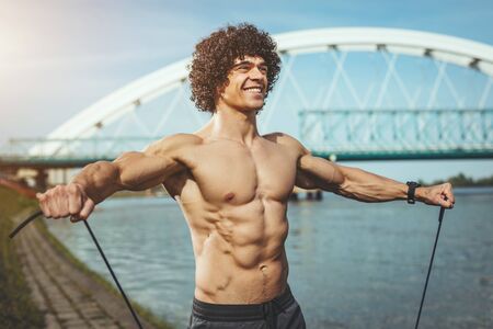 Fit muscular young man with naked torso doing exercise with resistance band near the river.の写真素材