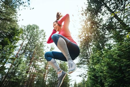 Young cheerful female runner jumping and enjoying life while jogging in a sunny forest.の写真素材