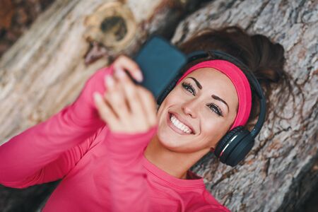 Young happy female runner lying on a wooden log and taking a break with music and smartphone after running in forest.の写真素材