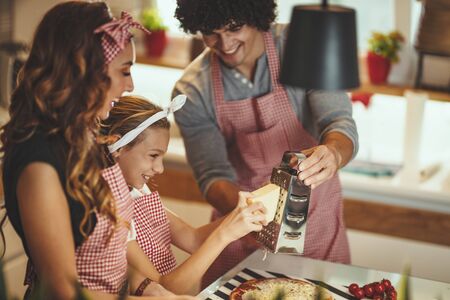 Happy parents and their daughter are preparing meal together in the kitchen. Little girl and her father are putting cheese on the pizza.の写真素材