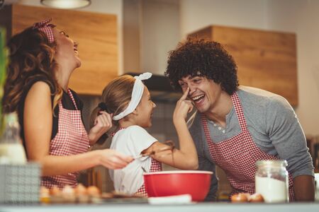 Happy parents and their daughter are preparing cookies together in the kitchen. Little girl is making fun putting some dough to mer father nose.の写真素材