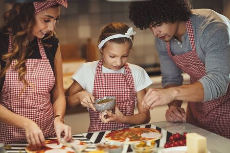 Happy parents and their daughter are preparing meal together in the kitchen while little girl is putting ketchup on the pizza dough.の写真素材