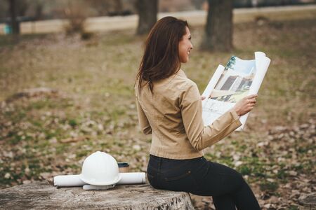 Successful young female engineer with white helmet beside her checked new project and analyzing blueprints in front of construction site.の写真素材
