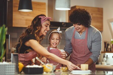 Happy parents and their daughter are preparing meal together in the kitchen while little girl is putting ketchup on the pizza dough.の写真素材