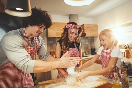 Happy parents and their daughter are preparing dough for pizza together in the kitchen. Little girl helps to her parents to mix dough on the table.の写真素材