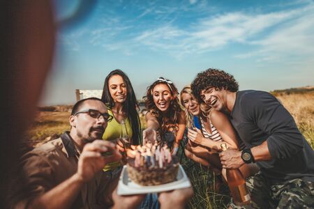 Young people have a good time in camp in nature. They're celebrating a birthday, laughing and greeting to their friend with birthday cake, happy to be together.の写真素材