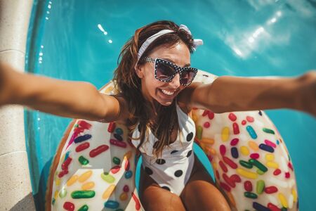 Beautiful young woman in swimming pool swims on inflatable ring donut and has fun making selfie in water on vacation.の写真素材