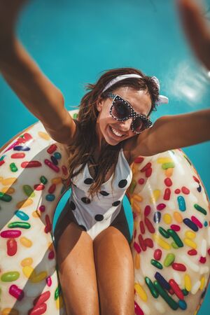 Beautiful young woman in swimming pool swims on inflatable ring donut and has fun making selfie in water on vacation.の写真素材
