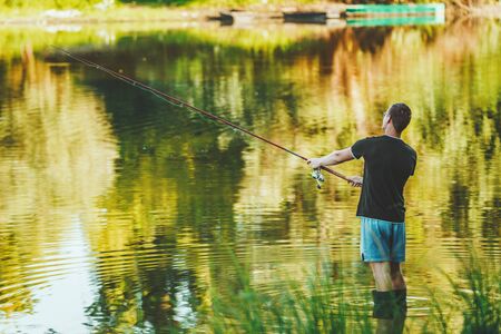 A young man in boots is fishing standing in shallow water freshwater of lake fringed with lush sceneryの写真素材