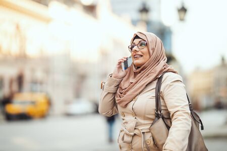Middle aged Muslim woman wearing hijab with a happy face standing in urban environment, taking on her smartphone.の写真素材