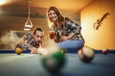 Young smiling cheerful couple is playing billiards in bar after work. They are having fun and involved in recreational activity.の写真素材