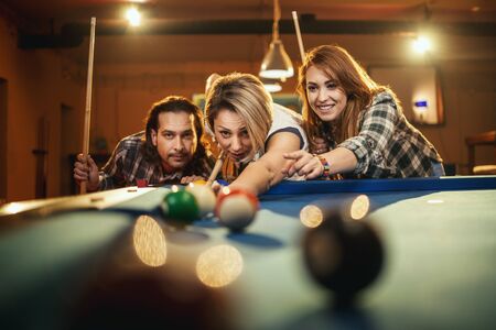 Young smiling cheerful friends are playing billiards in bar after work. They are involved in...