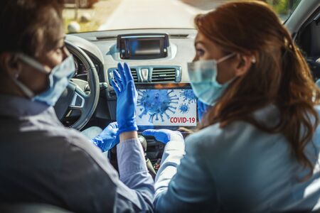 Young business women with protective masks on their faces are sitting in a car and talking about global crisis. They are showing each other an animation of a corona virus on a tablet.の写真素材