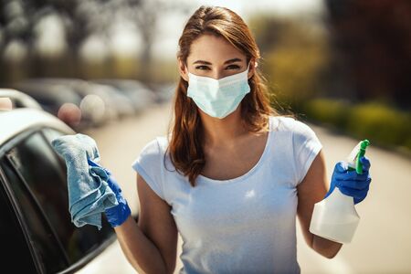 A young woman with a mask on her face and protective gloves on her hands is ready to wipe her car holding a cloth in one and a bottle with disinfectant in the other hand.の写真素材