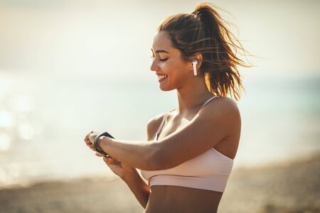 A beautiful young woman is looking at smartwatch and preparing to do jogging at the sea beach in summer sunny day.の写真素材