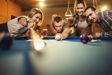 Group of a four smiling cheerful friends are playing billiards in bar after work. They are involved in recreational activity.の写真素材