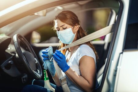 A young woman with a mask on her face and protective gloves on her nands wipes a car belt in her car...