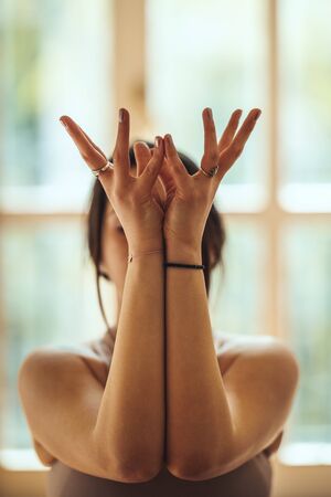 Young woman is doing yoga Lotus mudra during coronavirus pandemic in the living room at home. She is meditating in morning sunshine.の写真素材