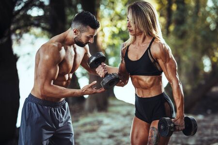 Attractive muscular female bodybuilder is doing hard exercises with personal trainer at the nature near the river.の写真素材