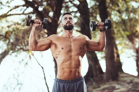 Fit muscular young man with naked torso is doing exercises with dumbbells in the nature near the...