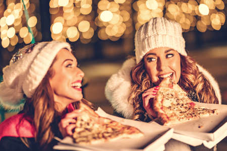 Two female friends enjoy the night out, looking each other and laughing with slices of pizza in their hands.の写真素材