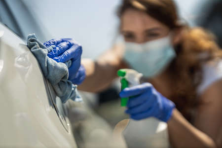 A young woman with a mask on her face and protective gloves on her hands, wipes her car holding a cloth in one and a bottle with disinfectant in the other hand.の写真素材