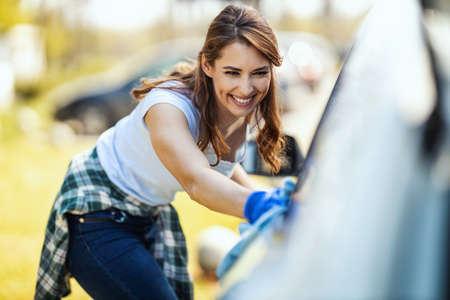 A smiling young woman with protective gloves on her nands wipes her car with a cloth ready to hit the road.の写真素材