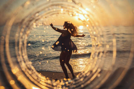 A beautiful creative composition of a sea landscape shot through a circle focus showing a young woman who is dancing on the beach in sunset.の写真素材