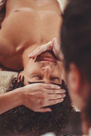 Handsome man is lying down on massage table and getting healthy facial massage by young female therapist in the spa center.の写真素材