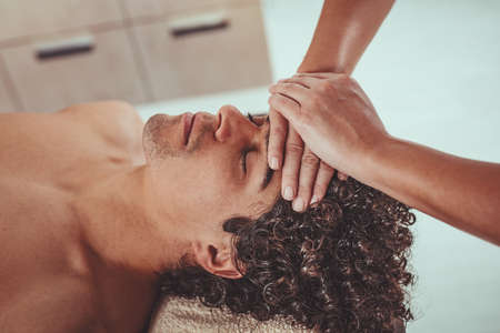 Handsome man is lying down on massage table and getting healthy facial massage by young female therapist in the spa center.の写真素材