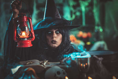 Witch with awfully face in creepy surroundings and smoky green background talks magic words to bones above boiling cauldron. Halloween concept.の写真素材