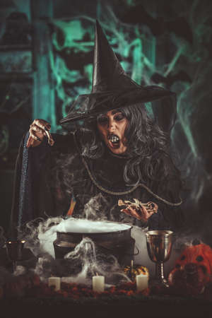 Witch with awfully face in creepy surroundings and smoky green background talks magic words to bones above boiling cauldron. Halloween concept.の写真素材
