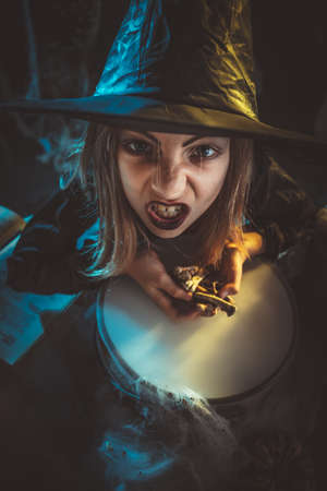 Young angry witch with awfully face tells evil words, and putting bones in cauldron with poison...