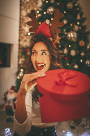A surprised young woman is sitting on the floor beside Christmas tree and holding the magic present in the red box she just received.の写真素材