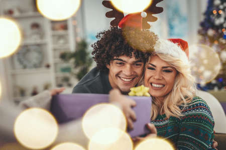 Loving couple dressed in cozy warm sweaters is spending time in the living room on Christmas - New Year eve. Young man surprises his girlfriend giving her a gift box.の写真素材