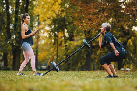 A mature muscular woman is doing squat exercises with personal trainer in the park, dressed in a black suit with an EMS electronic simulator to stimulate her muscles.の写真素材