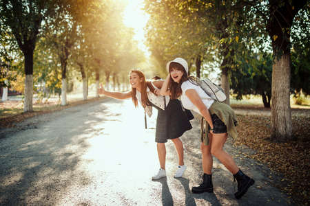 Two beautiful young smiling women with backpacks on their back are walking along the autumn sunny avenue and hitchhiking.の写真素材