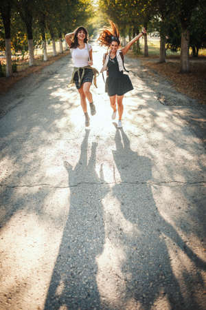 Two beautiful cheerful young women with backpacks on their back are walking along the autumn sunny avenue and holding hands up.の写真素材