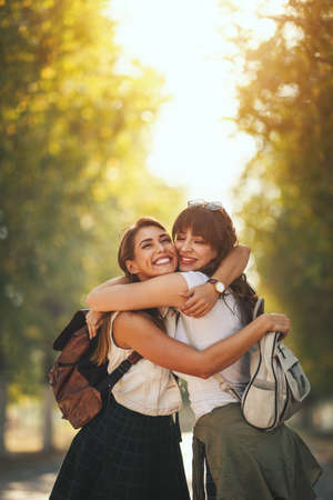 Two beautiful young smiling women with backpacks on their back are walking along the autumn sunny avenue and hugging.の写真素材