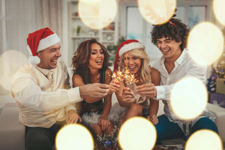 Two young cheerful couples are celebrating New Year at home. They are having fun with sparkles in...