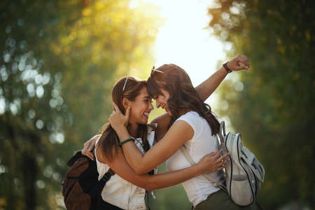 Two beautiful young smiling women with backpacks on their back are walking along the autumn sunny avenue and hugging.の写真素材