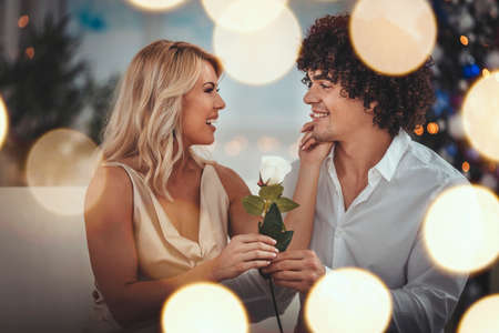 Beautiful young couple is celebrating Christmas at home. Handsome smiling man is giving a white rose...