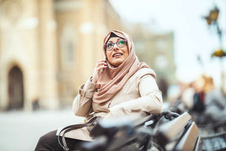 Middle aged Muslim woman wearing hijab with a happy face is sitting on the bench in urban environment, talkings on her smartphone.の写真素材
