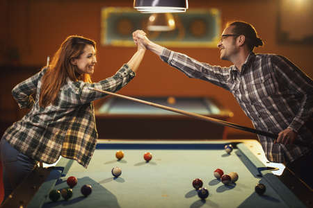 Young smiling cheerful couple is playing billiards in bar after work. They are having fun and celebrate victory.の写真素材