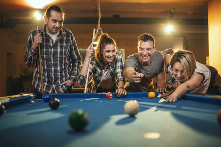 Young smiling cheerful friends are playing billiards in bar after work.  They are involved in...