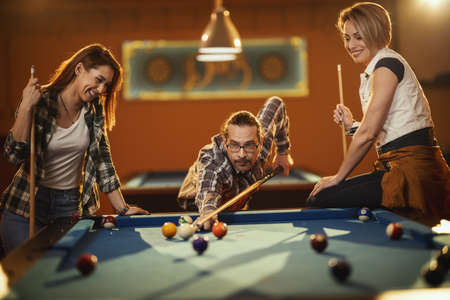 Young smiling cheerful friends are playing billiards in bar after work. They are involved in...