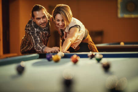 Young smiling cheerful couple is playing billiards in bar after work. They are having fun and ...